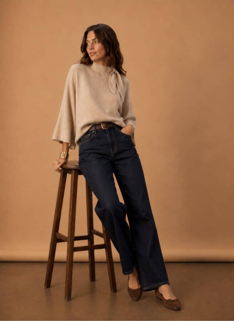 Stylish woman in oatmeal cape knit and dark jeans against beige background.