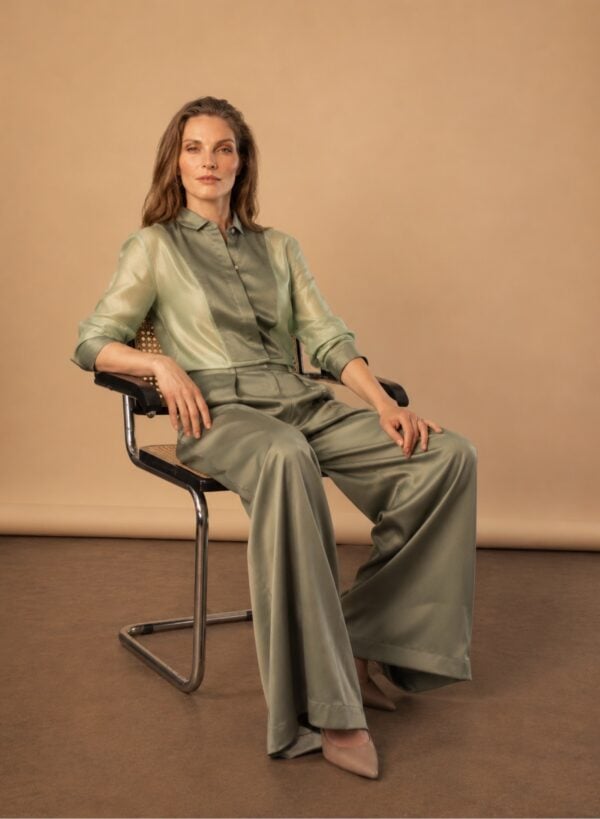 Elegant woman in satin tuxedo and wide-leg trousers, seated on a modern chair.