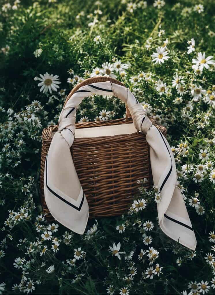 Woven basket bag with a silk scarf handle on daisies in a garden.