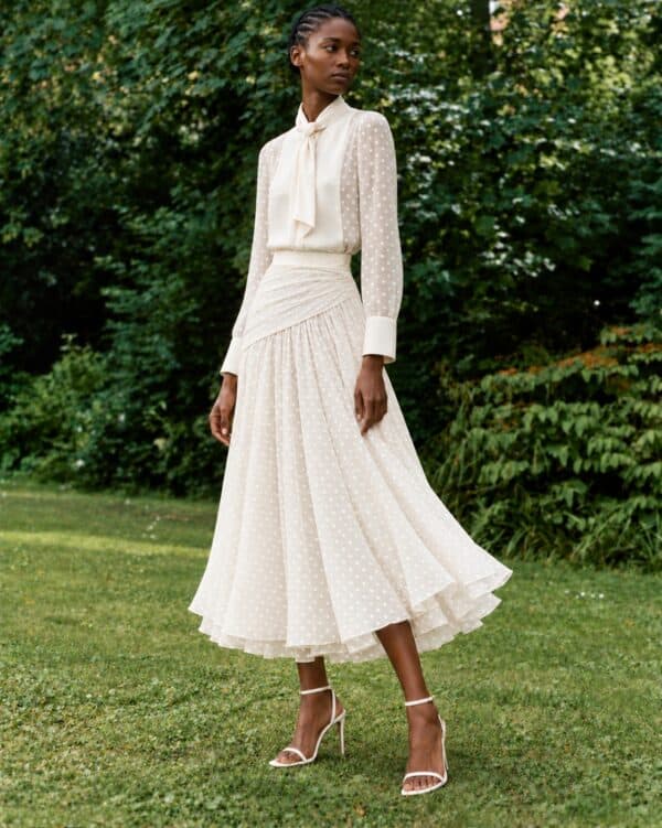 Front three-quarter garden view of the white Gilda shirt and skirt, with a polka-dot cotton yoke and white strappy heels.