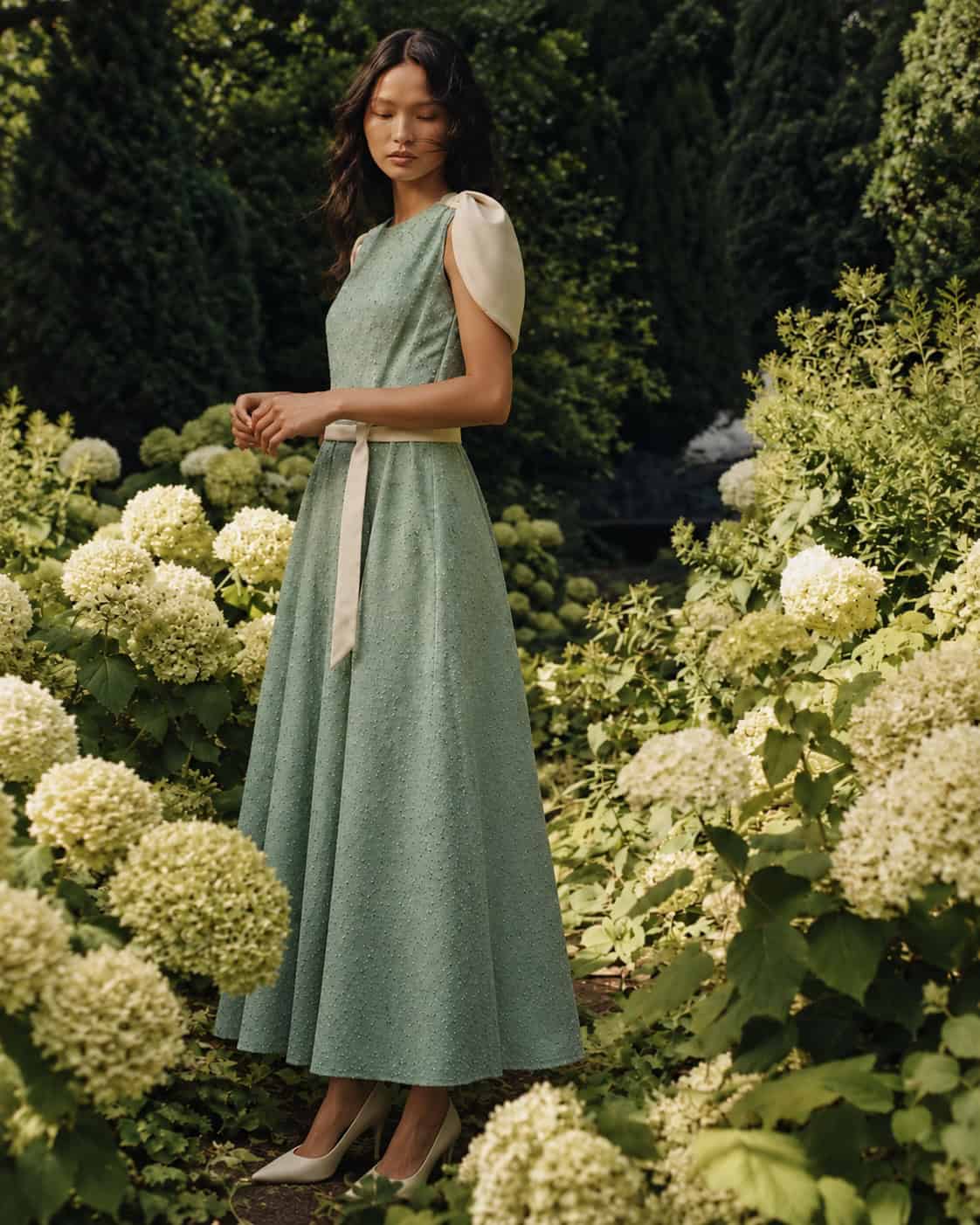 Full-length sage green Catherine dress with ivory satin details and pointed heels among green hydrangeas